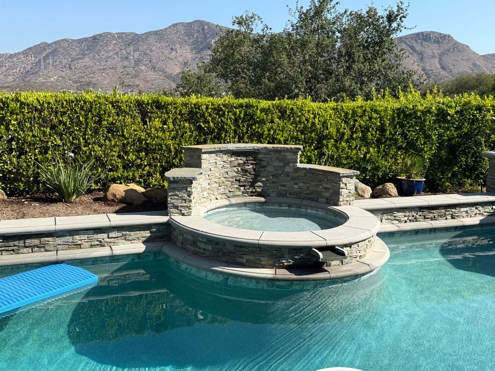 Stone hot tub beside a serene pool with mountains and greenery in the background.